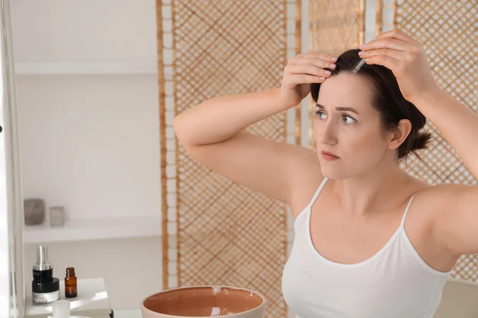 Žena si při zrcadle nanáší sérum ke kořínkům vlasů