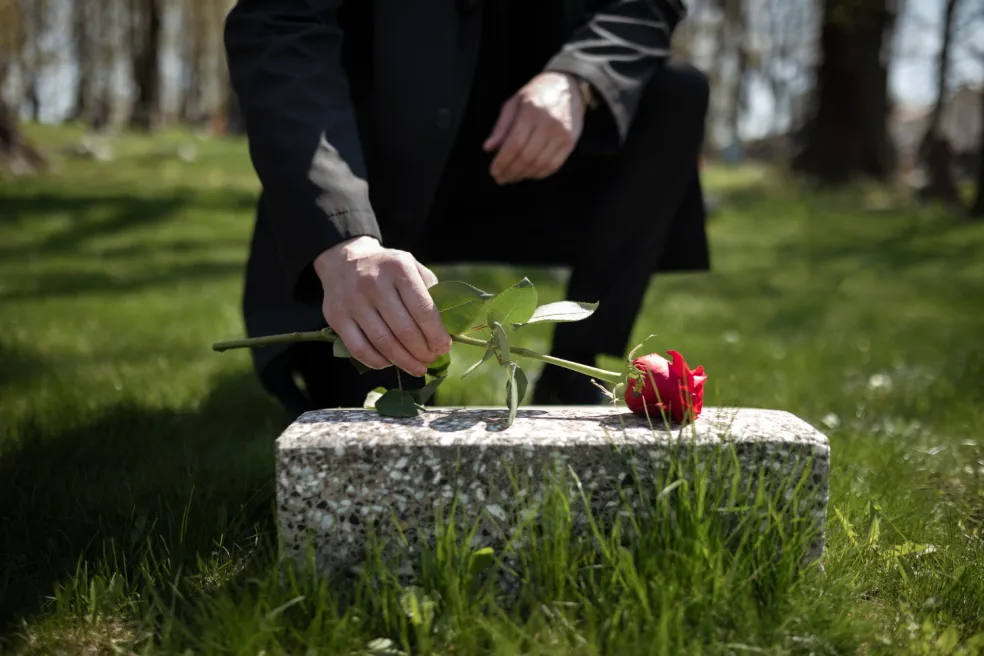 Muž v černém pokládá červenou růži na hrob na hřbitově