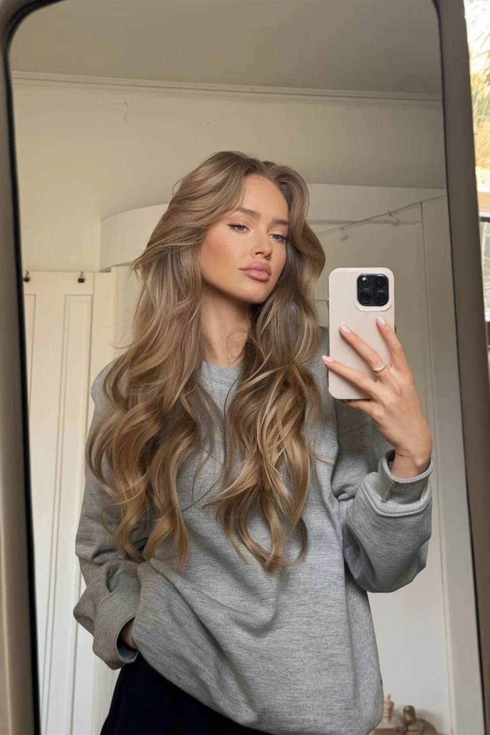 Žena s dlouhými vlnitými vlasy v béžovo-hnědém odstínu s jemnými blond odlesky si fotí selfie v zrcadle